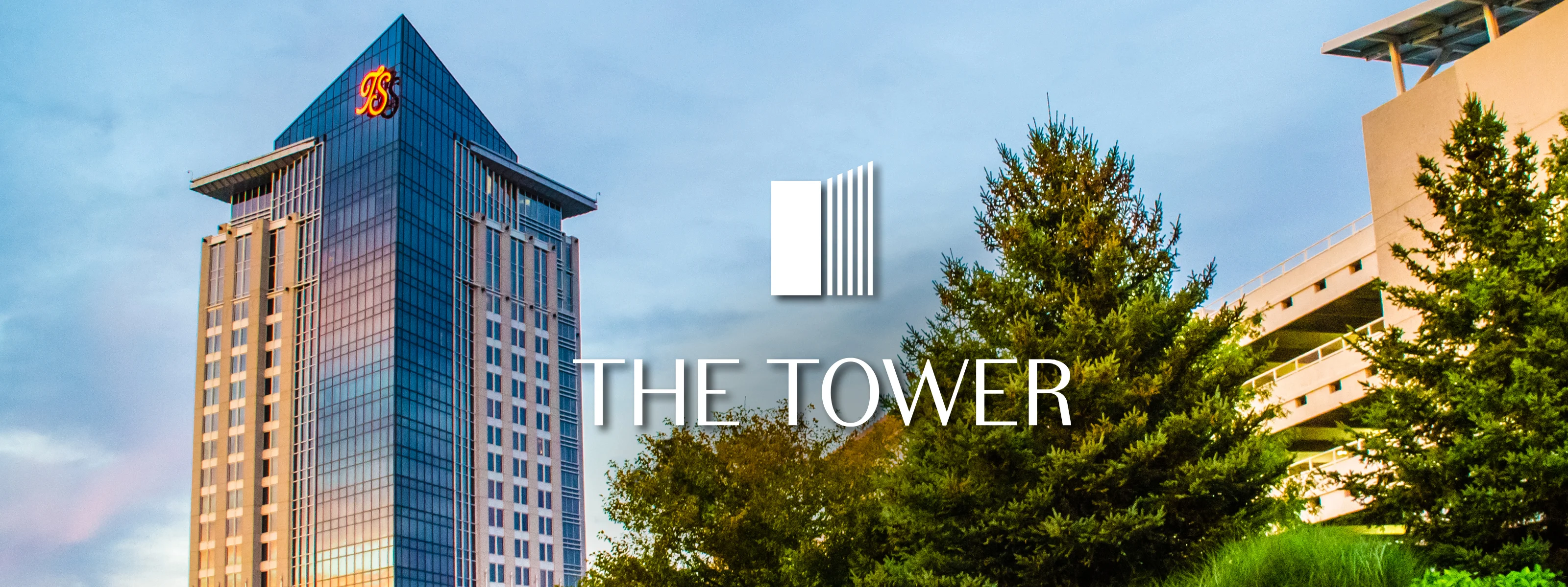 Trees and flowers line the approach to The Tower at Turning Stone. The setting sun illuminates the glass on The Tower.